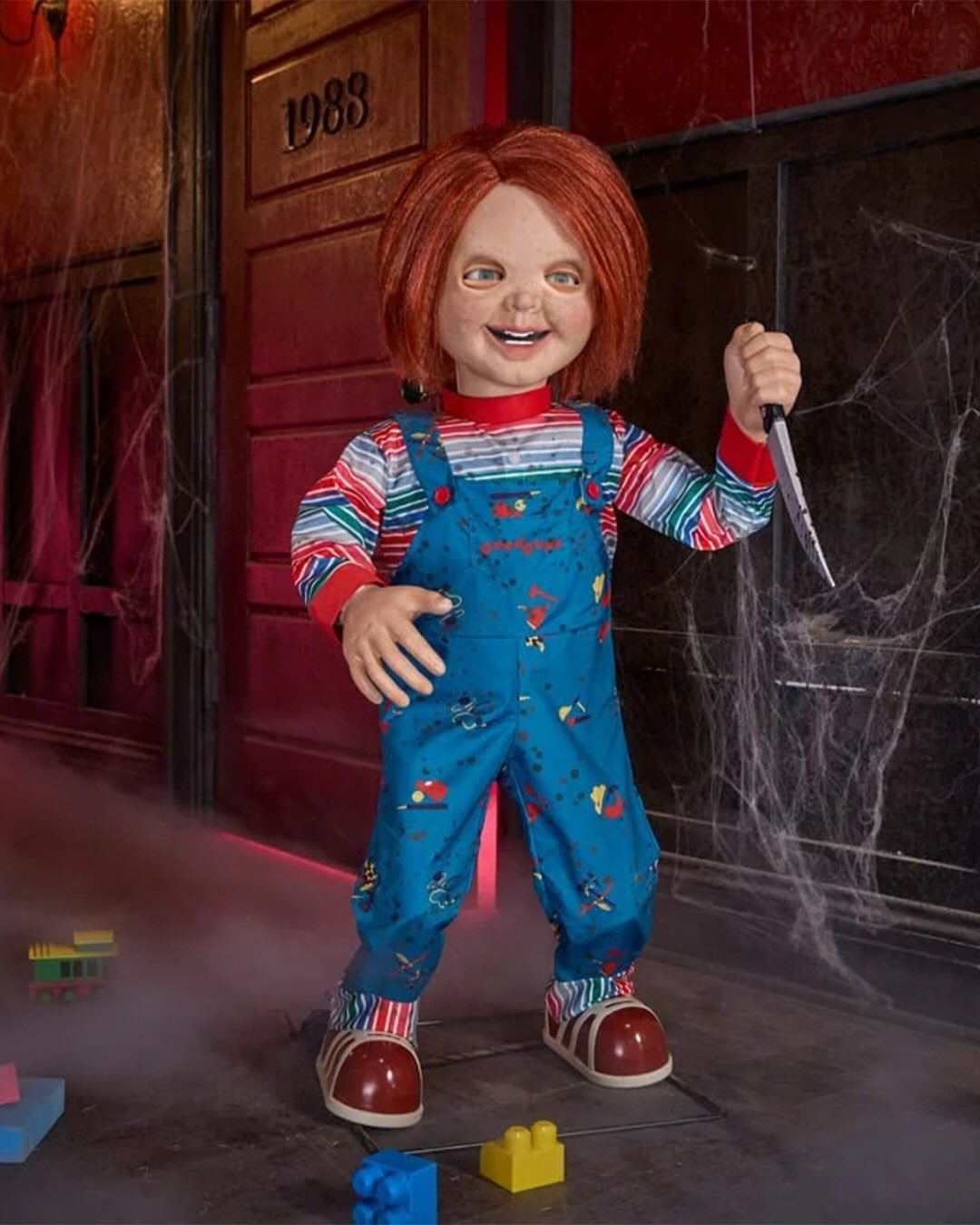 Life-size Chucky animatronic in a striped shirt and dungarees holding a knife outside a cobwebbed doorway